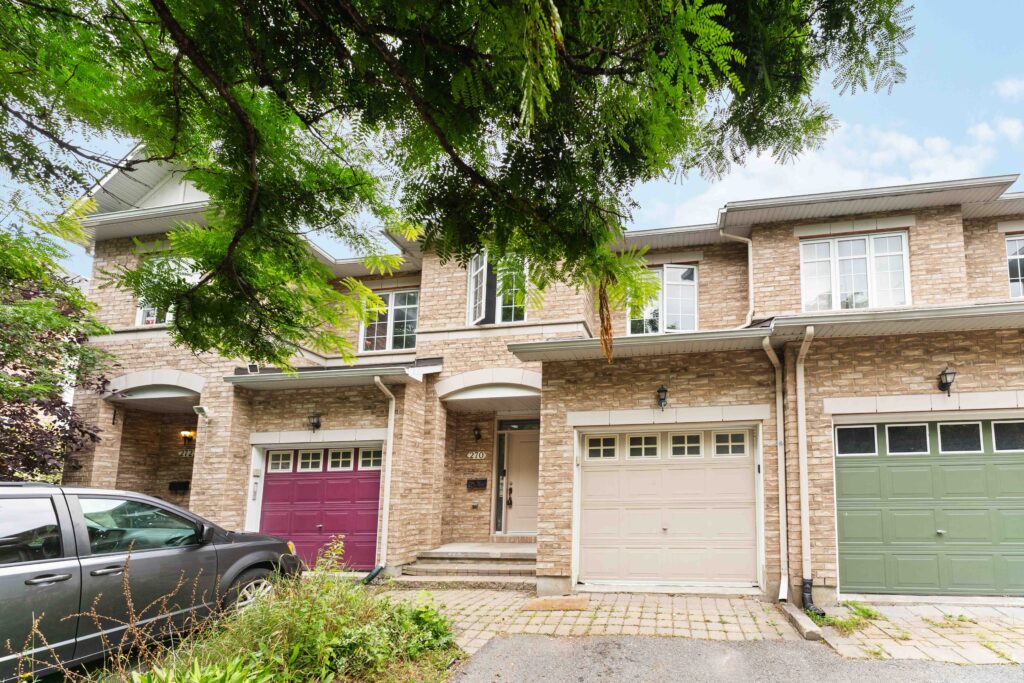 Front exterior of a 3 bedroom Ottawa rental property townhouse featuring a brick facade, single car garage, private driveway, and tree lined residential street in a family friendly neighbourhood. Anna Alemi Real Estate Team. Best Ottawa Realtor.